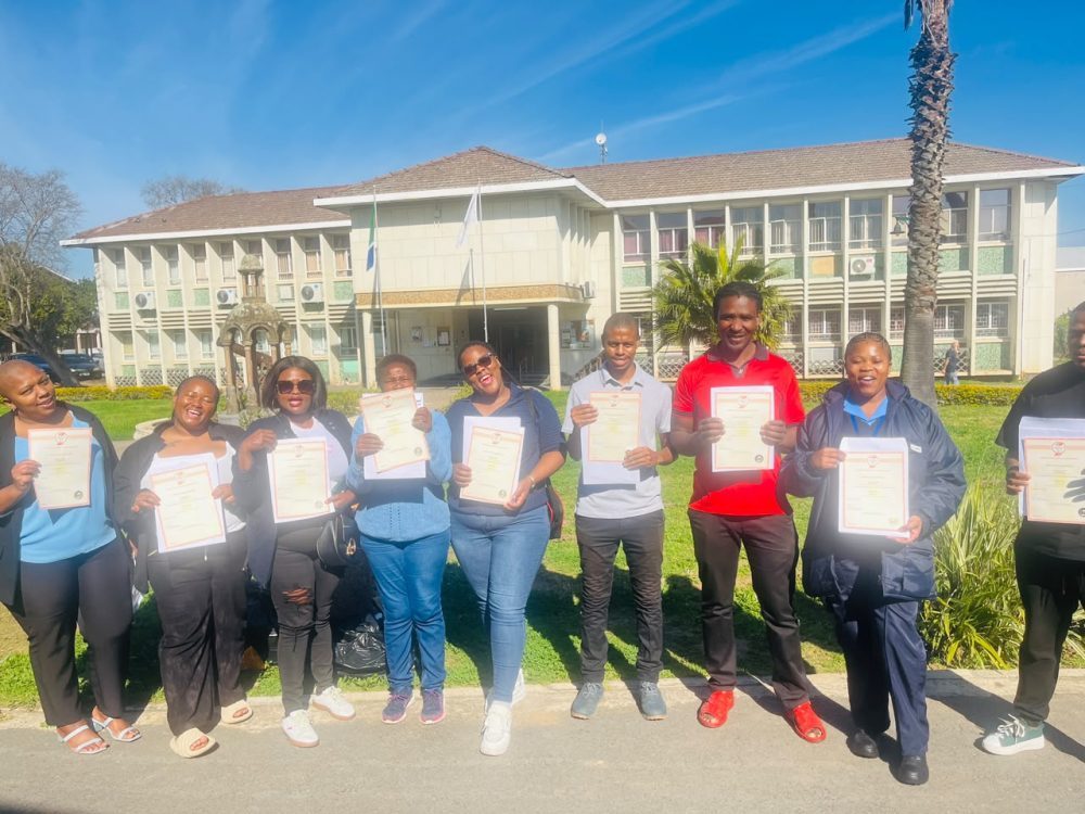 Learners holding certificates of competence outside a public-sector building after completing an OMEIDA skills programme